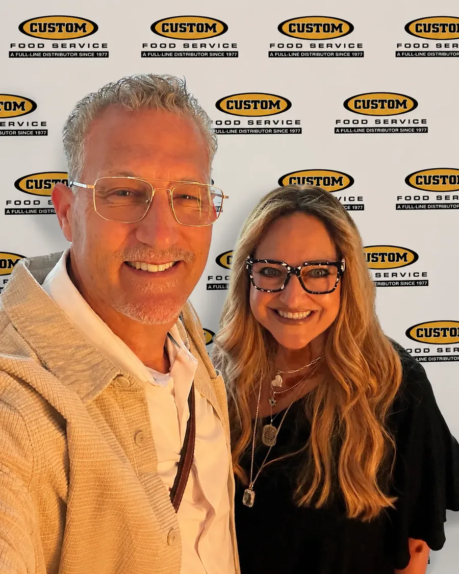 David and Linda Schnitzer, second-generation owners of Custom Food Service, smiling in front of a Custom Food Service event backdrop
