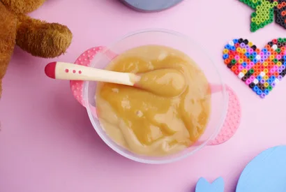 Creamy applesauce or fruit puree in a white bowl with pink handles and infant feeding spoon, surrounded by toys on pink background.