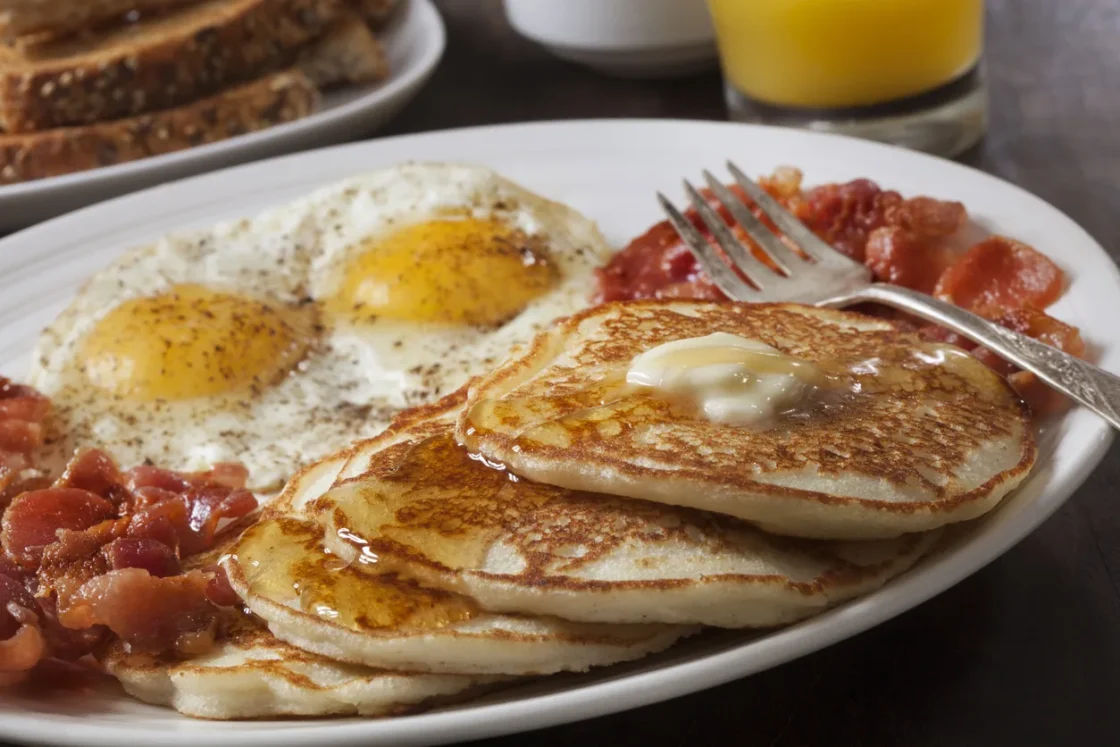 A complete breakfast plate featuring golden pancakes with butter, sunny-side up eggs, and crispy bacon strips, ideal for foodservice and restaurant supply.