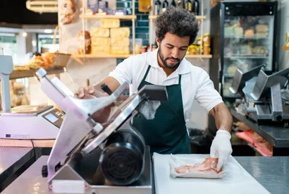 A skilled butcher in apron uses a commercial meat slicer to prepare fresh meat portions in a professional food service kitchen environment.