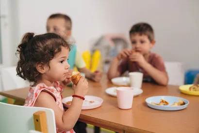 Young children eating healthy meals at a table in a cafeteria or institutional food service setting, demonstrating proper nutrition and food service standards for youth programs.