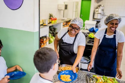 Commercial foodservice workers in aprons and hairnets preparing plated meals in a professional kitchen, demonstrating wholesale food preparation and food service industry operations.