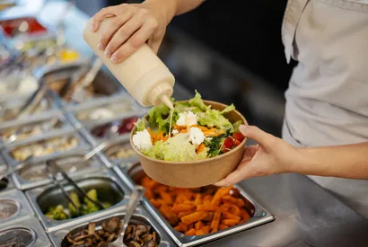 Food service worker preparing a fresh garden salad bowl with mixed vegetables and toppings from ingredient bins in commercial kitchen setup.
