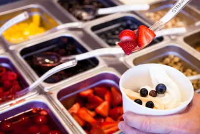 Commercial frozen yogurt dispensing station with multiple ingredient bins including fresh strawberries, blueberries, and colorful fruit toppings for wholesale food service operations.