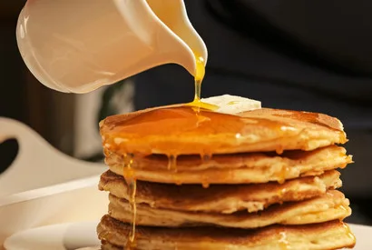Stack of golden pancakes with maple syrup being poured over top, ideal for food service and commercial food industry applications.