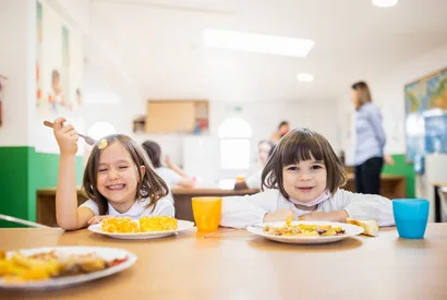 Two smiling children eating healthy meals at a school cafeteria table, demonstrating quality food service for institutional dining and student nutrition programs.